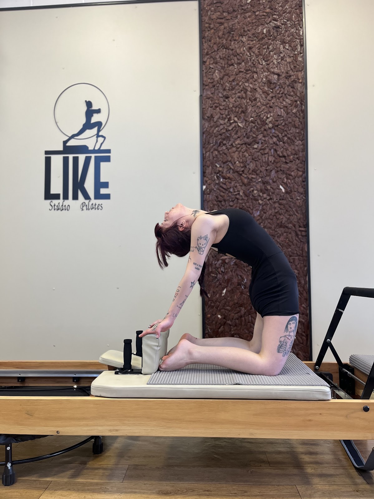 Aluna da Like Pilates em exercício de extensão sobre o reformer, ao lado da parede com a marca do estúdio